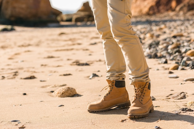 Gambe in stivali calzature di un giovane hipster che viaggia con uno zaino sulla costa del mare autunnale indossando giacca e cappello caldi