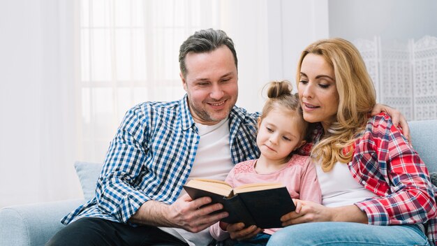 Genitore che si siede con la figlia che si siede sul libro di lettura del sofà