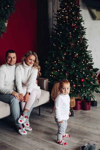 Genitori amorevoli in vestiti bianchi che si siedono sul divano vicino all'albero di Natale mentre guardano il loro bambino