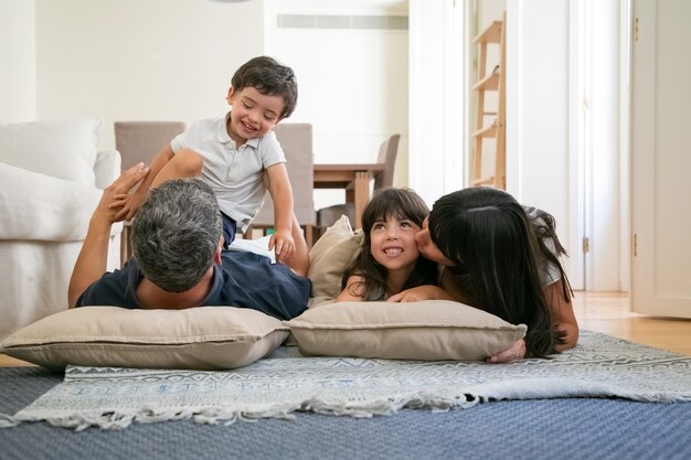 Genitori divertenti allegri che stringono a sé, abbracciano e baciano i bambini piccoli, divertendosi insieme