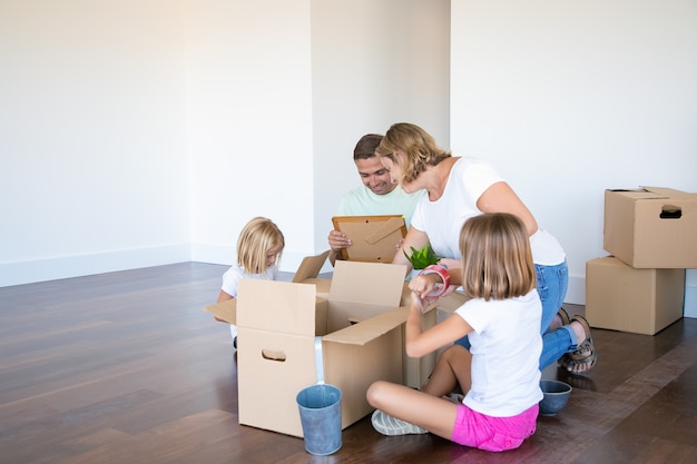 Genitori e bambini felici che disimballano le cose nel nuovo appartamento vuoto, seduti sul pavimento e prendono oggetti dalle scatole aperte