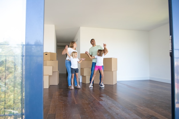 Genitori e figlie allegri che ballano e si divertono vicino a un mucchio di scatole mentre si trasferiscono nel nuovo appartamento