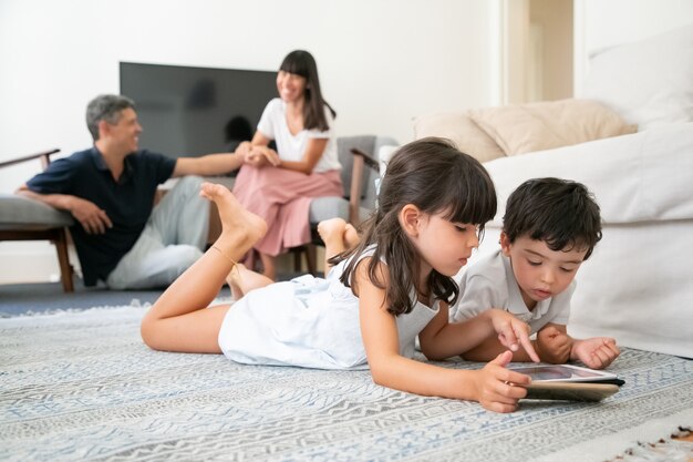 Genitori felici che guardano bambini carini sdraiati sul pavimento in soggiorno e utilizzano gadget digitali con app di apprendimento.