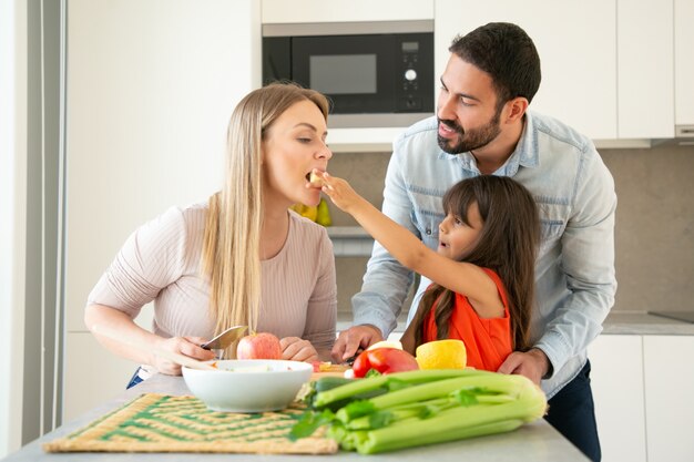 Genitori felici e bambino che si divertono a cucinare insieme. Ragazza che dà una fetta di verdura alla mamma per il gusto mentre la mamma taglia frutta e verdura fresca. Cucina familiare o concetto di stile di vita