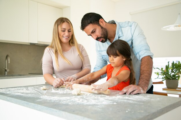 Genitori positivi guardando la figlia che rotola pasta sulla scrivania della cucina con farina disordinata. Giovani coppie e la loro ragazza che cuociono insieme i panini o le torte. Concetto di cucina familiare