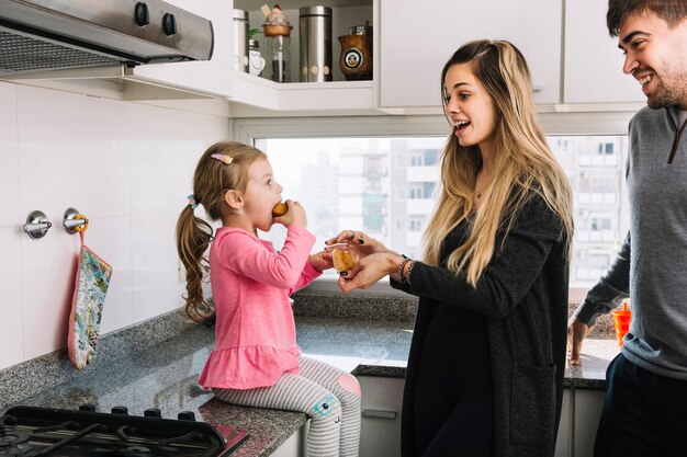 Genitori sorridenti che esaminano la loro figlia che mangia bigné in cucina