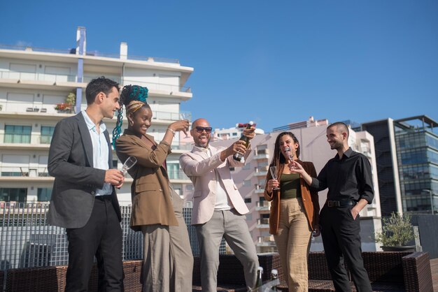 Gente di affari allegra sulla festa sul tetto della terrazza. Uomini e donne in abiti formali in piedi sul tetto della terrazza, aprendo champagne, ridendo. Teambuilding, concetto di festa