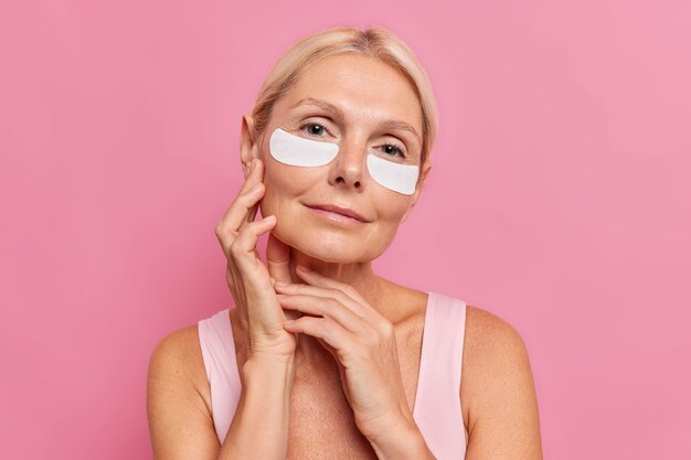 Gentile donna di mezza età con i capelli biondi tocca il viso applica teneramente cerotti di bellezza sotto gli occhi per ridurre le rughe indossa un trucco minimo vestito con una maglietta ha una pelle sana isolata sul muro rosa