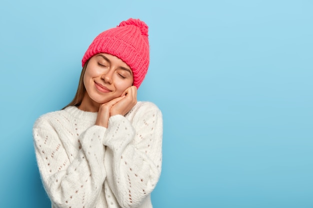 gentile giovane donna chiude gli occhi, inclina la testa, immagina qualcosa di piacevole, indossa un cappello rosa, maglione invernale, posa su sfondo blu