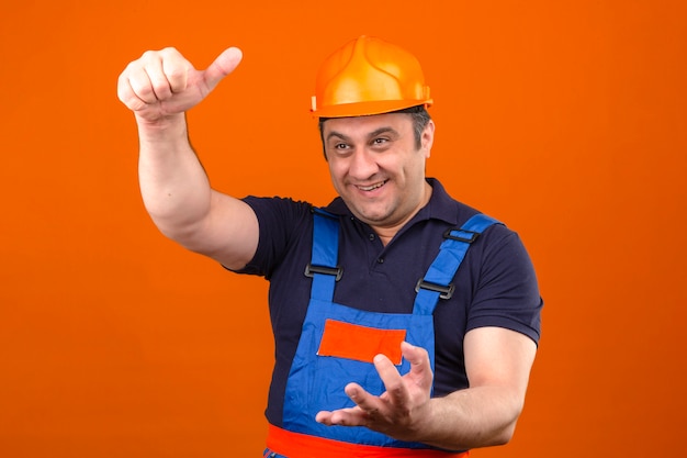 Gestore d'uso dell'uomo uniforme del costruttore e casco di sicurezza sorridenti che gesturing mostrando i pollici su sopra la parete arancio isolata