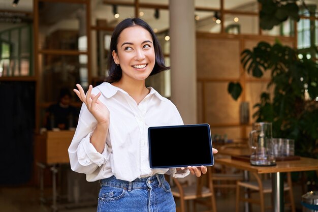 Gestore del caffè della donna asiatica sorridente amichevole che mostra lo schermo del tablet e che sembra felice dimostrando il suo profitto del ristorante