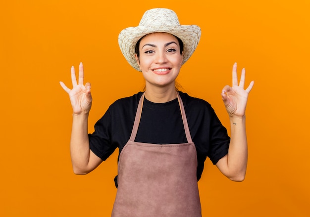 Giardiniere della giovane bella donna in grembiule e cappello che esaminano sorridere anteriore che mostra segno giusto che sta sopra la parete arancione