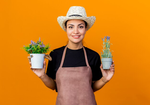 Giardiniere della giovane bella donna in grembiule e cappello che tengono le piante in vaso che esaminano davanti che sorride con la faccia felice che sta sopra la parete arancione