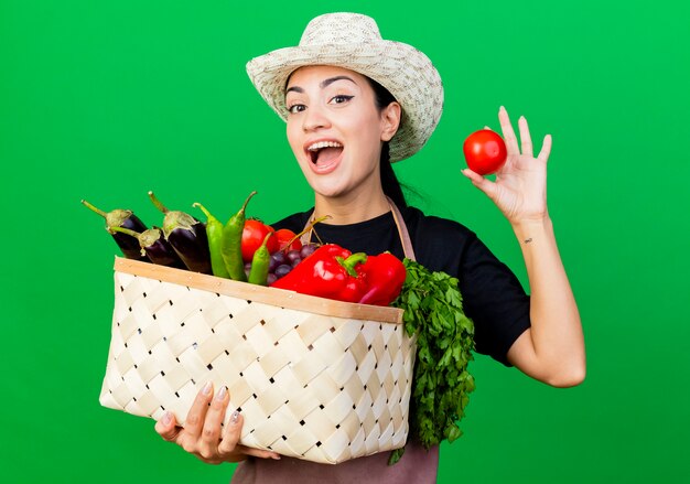 Giardiniere della giovane bella donna in grembiule e cappello che tiene il cestino pieno di verdure che mostrano pomodoro felice ed eccitato in piedi sopra la parete verde