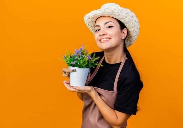 Giardiniere della giovane bella donna in grembiule e cappello che tiene la pianta in vaso che esamina sorridente anteriore che sta sopra la parete arancione