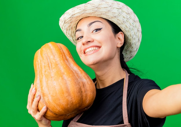 Giardiniere della giovane bella donna in grembiule e cappello che tiene la zucca facendo selfie felice ed emozionato