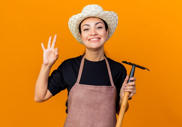 Giardiniere della giovane bella donna in grembiule e cappello che tiene mini rastrello guardando davanti sorridente che mostra segno giusto in piedi sopra la parete arancione