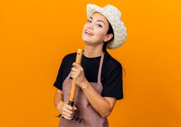 Giardiniere della giovane bella donna in grembiule e cappello che tiene mini rastrello guardando davanti sorridente con la faccia felice in piedi sopra la parete arancione