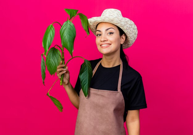 Giardiniere della giovane bella donna in grembiule e cappello che tiene pianta guardando la parte anteriore sorridente con la faccia felice in piedi sopra la parete rosa