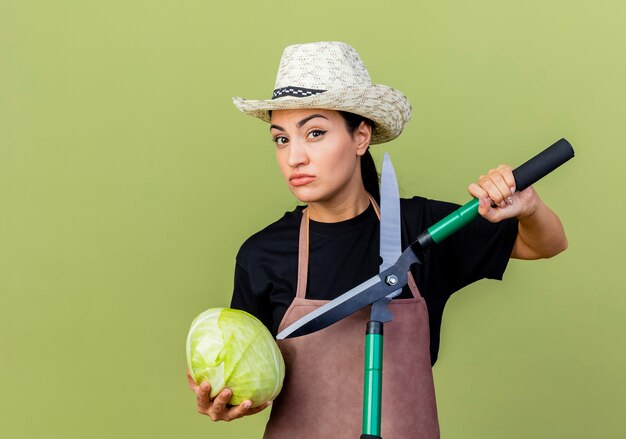 Giardiniere di giovane bella donna in grembiule e cappello che tiene cavolo e tagliasiepi guardando davanti con faccia seria in piedi sopra il muro verde chiaro