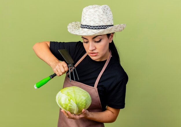 Giardiniere di giovane bella donna in grembiule e cappello che tiene cavolo e vanga guardandolo con faccia seria in piedi sopra la parete verde chiaro