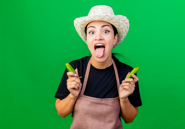 Giardiniere di giovane bella donna in grembiule e cappello che tiene peperoncino verde rotto che attacca fuori la lingua che sta sopra fondo verde