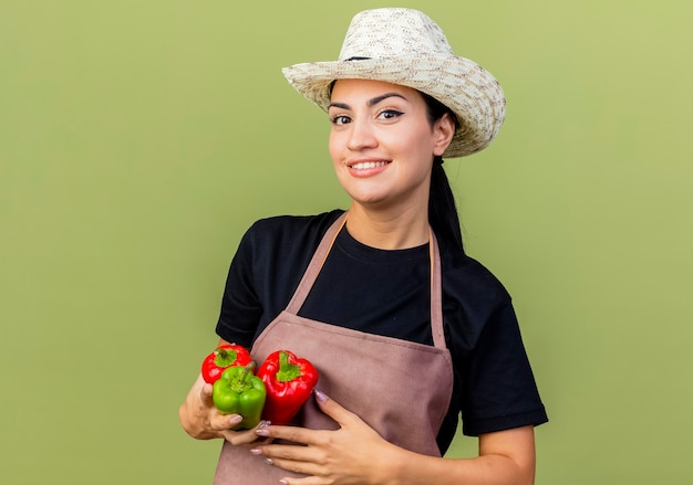 Giardiniere di giovane bella donna in grembiule e cappello che tiene peperoni colorati guardando davanti sorridente allegramente in piedi sopra il muro verde chiaro