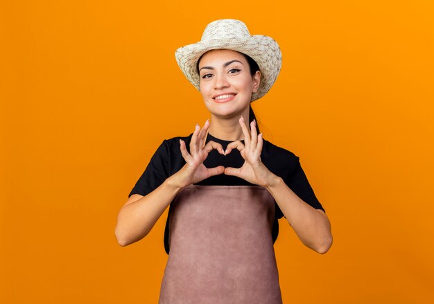 Giardiniere di giovane bella donna in grembiule e cappello guardando davanti facendo il gesto del cuore con le dita sorridenti in piedi sopra la parete arancione