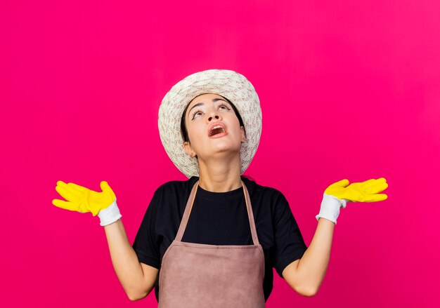 Giardiniere di giovane bella donna in guanti di gomma grembiule e cappello che osserva in su confuso diffondendo le braccia ai lati in piedi sopra il muro rosa