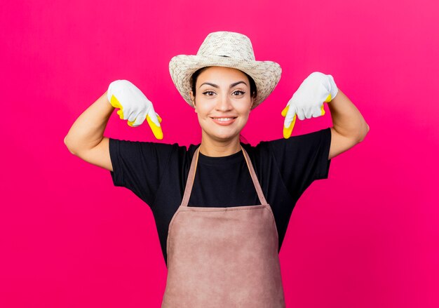 Giardiniere di giovane bella donna in guanti di gomma grembiule e cappello che punta con il dito indice verso il basso sorridente in piedi sopra il muro rosa