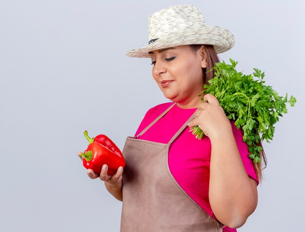 Giardiniere di mezza età donna in grembiule e cappello azienda peperone rosso fresco ed erbe guardando il pepe con il sorriso sul viso in piedi su sfondo bianco