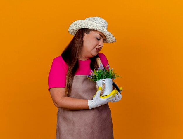 Giardiniere di mezza età donna in grembiule e cappello tenendo la pala e pianta in vaso guardando la pianta con il sorriso sul viso in piedi su sfondo arancione