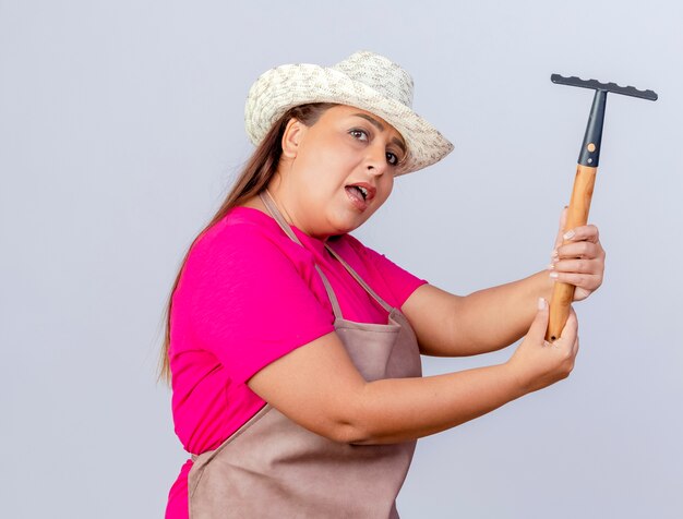 Giardiniere donna di mezza età in grembiule e cappello oscillante mini rastrello che guarda l'obbiettivo con il viso serio in piedi su sfondo bianco