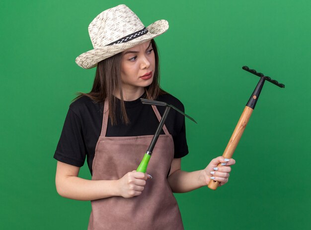 Giardiniere femmina caucasica piuttosto incapace che indossa un cappello da giardinaggio che tiene il rastrello della zappa e guarda il rastrello