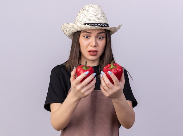 Giardiniere femminile abbastanza caucasico ansioso che porta la tenuta del cappello di giardinaggio e che esamina i peperoni rossi su bianco