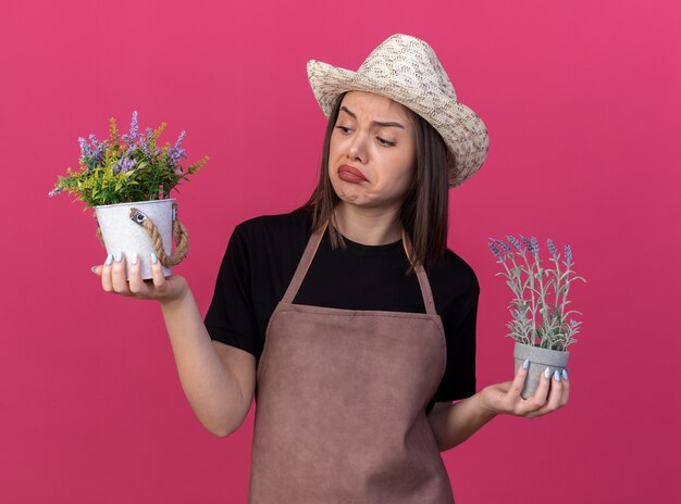 Giardiniere femminile abbastanza caucasico impressionato che indossa il cappello di giardinaggio che tiene e che esamina i vasi da fiori isolati sulla parete rosa con lo spazio della copia