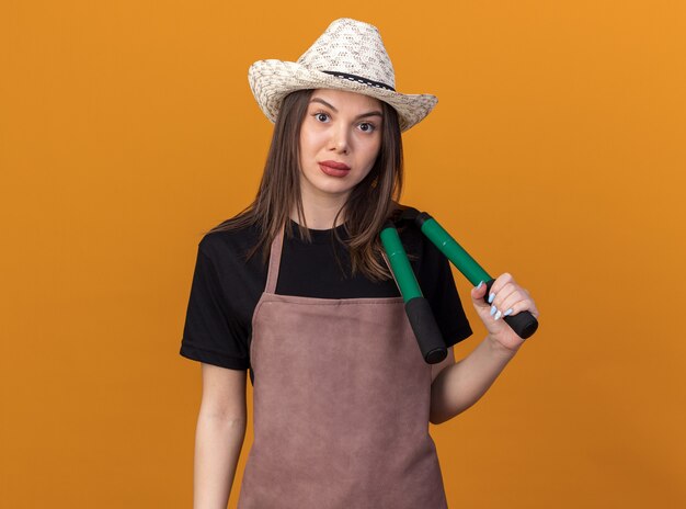 Giardiniere femminile abbastanza caucasico sicuro che porta il cappello di giardinaggio che tiene le forbici di giardinaggio sulla spalla sull'arancia