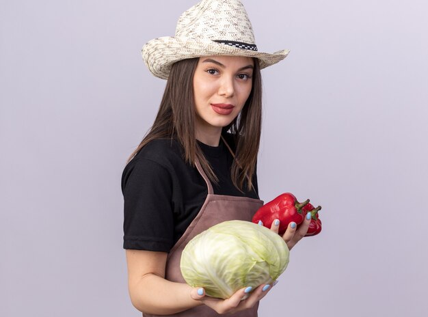 Giardiniere femminile abbastanza caucasico sicuro che porta il cappello di giardinaggio che tiene peperoni rossi e cavolo su bianco