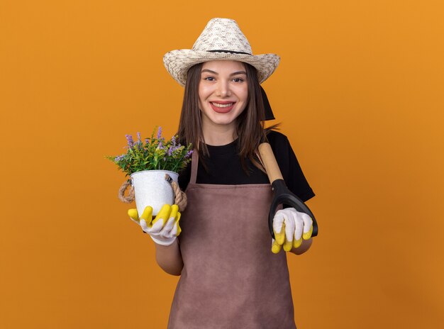 Giardiniere femminile abbastanza caucasico sorridente che indossa cappello e guanti di giardinaggio che tengono vaso di fiori e vanga sulla spalla sull'arancio
