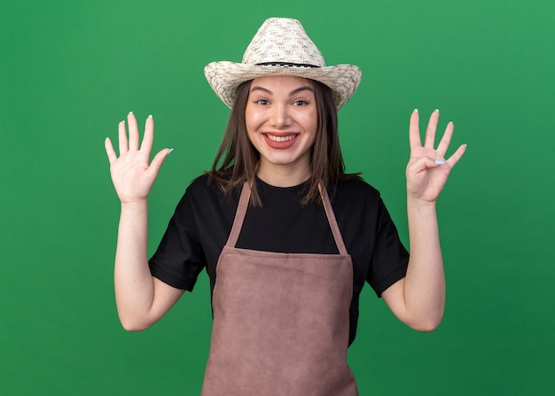 Giardiniere femminile abbastanza caucasico sorridente che porta il cappello di giardinaggio che gesturing nove con le dita sul verde