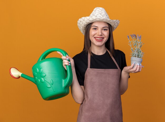 Giardiniere femminile abbastanza caucasico sorridente che porta il cappello di giardinaggio che tiene annaffiatoio e vaso di fiori sull'arancia