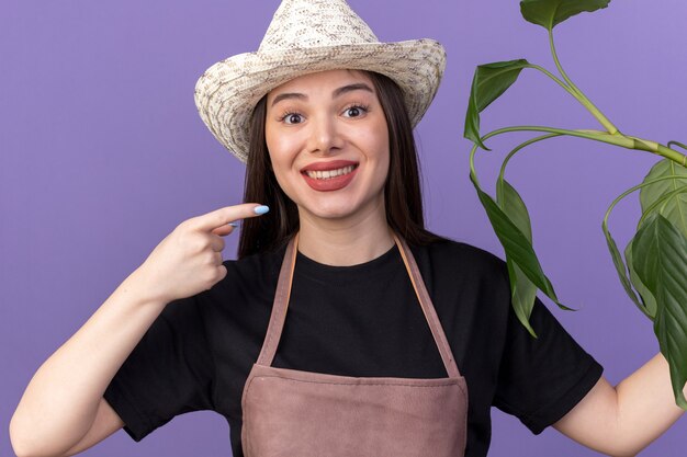 Giardiniere femminile abbastanza caucasico sorridente che porta la tenuta del cappello di giardinaggio e che indica al ramo della pianta isolato sulla parete viola con lo spazio della copia