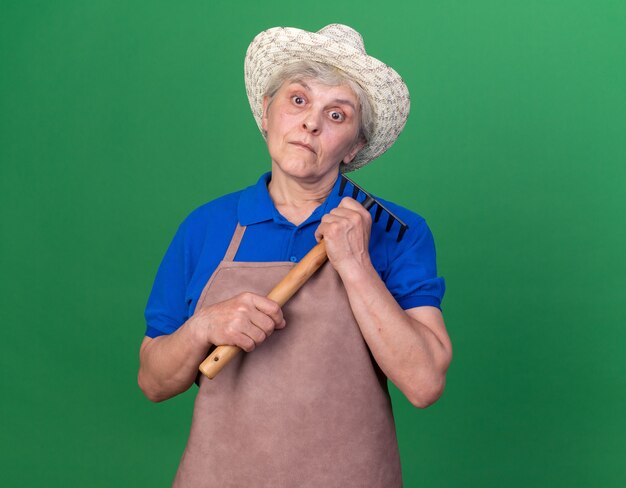 Giardiniere femminile anziano scioccato che indossa il cappello di giardinaggio che tiene il rastrello sul verde