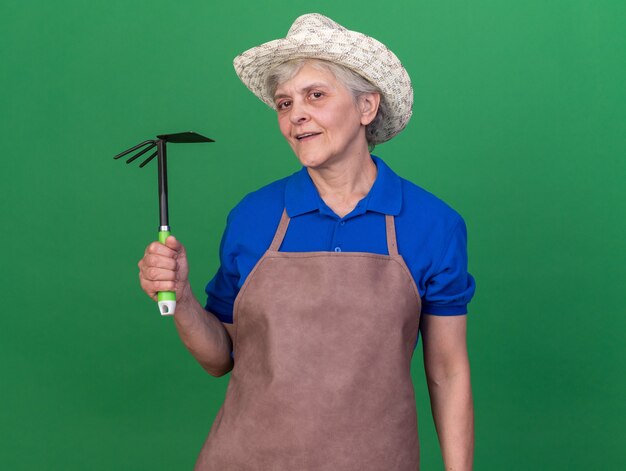 Giardiniere femminile anziano sicuro che porta il cappello di giardinaggio che tiene il rastrello della zappa sul verde