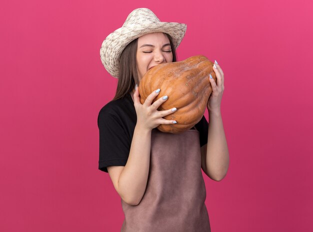 Giardiniere femminile caucasico abbastanza affamato che indossa cappello da giardinaggio che finge di mordere la zucca isolata sulla parete rosa con lo spazio della copia