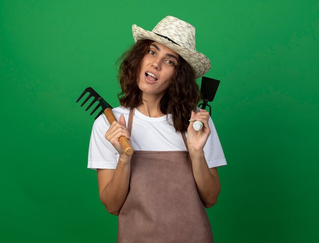 Giardiniere femminile giovane gioioso in cappello da giardinaggio da portare uniforme che mette il rastrello con il rastrello della zappa sulle spalle