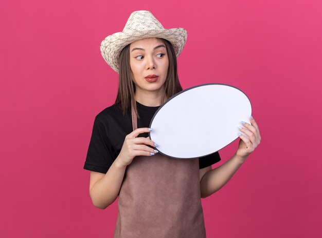 Giardiniere femminile piuttosto caucasico ansioso che indossa il cappello di giardinaggio che tiene il fumetto e che esamina il lato sul colore rosa