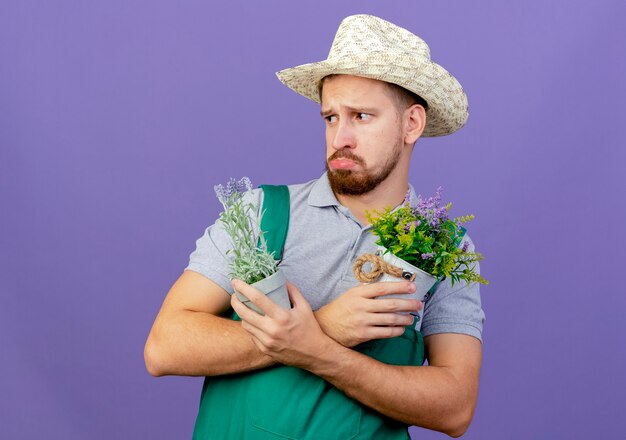 Giardiniere slavo bello giovane triste in uniforme e cappello tenendo le mani incrociate tenendo i vasi da fiori guardando il lato isolato sulla parete viola con lo spazio della copia