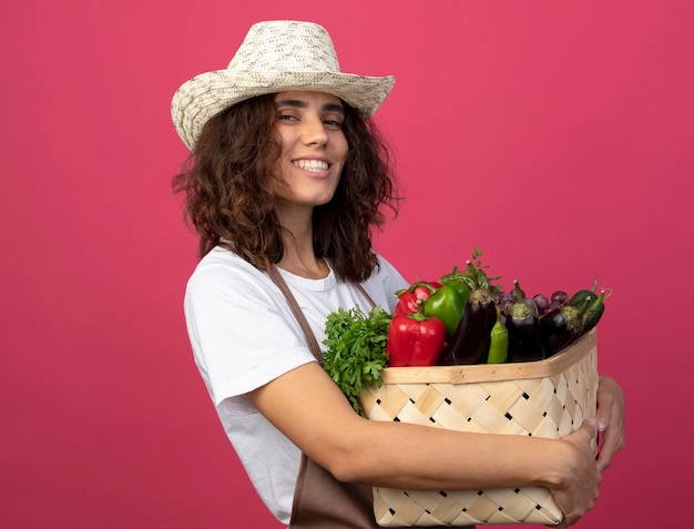 Giardiniere sorridente della giovane donna in uniforme che porta il cappello di giardinaggio che tiene il cestino della verdura isolato sul colore rosa