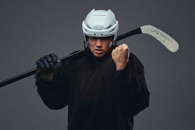 Giocatore di hockey professionista arrabbiato in abiti sportivi neri in piedi con una mazza da hockey. Isolato su sfondo grigio.